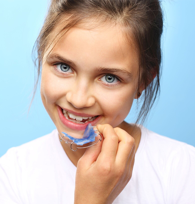 Young girl holding colorful orthodontic braces, smiling against blue background for dental practice.