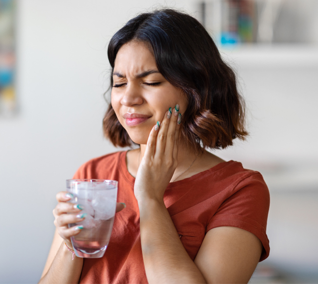 Woman in red shirt holding water glass, touching jaw in pain, home setting.