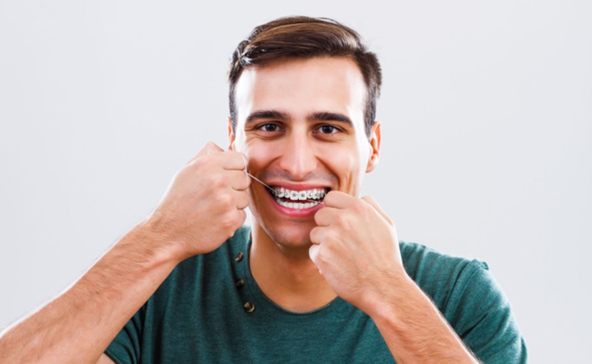 Young man in teal shirt smiling, pointing to teeth against white background for orthodontics website.