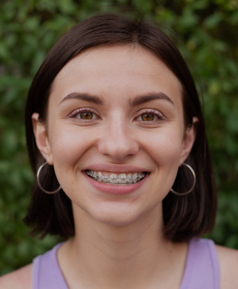 Young woman smiling at camera wearing braces, portrait style headshot with green foliage background.