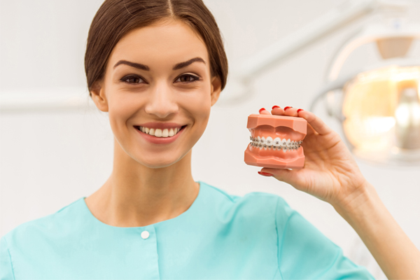 Dental professional in turquoise scrubs holding orthodontic model with braces in clinical setting.