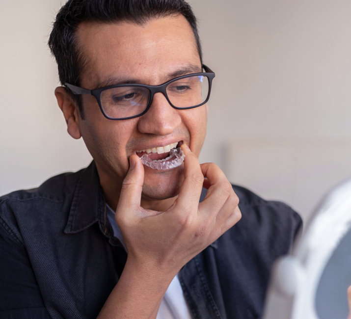 Young man wearing glasses smiling while touching his teeth in dental office setting.