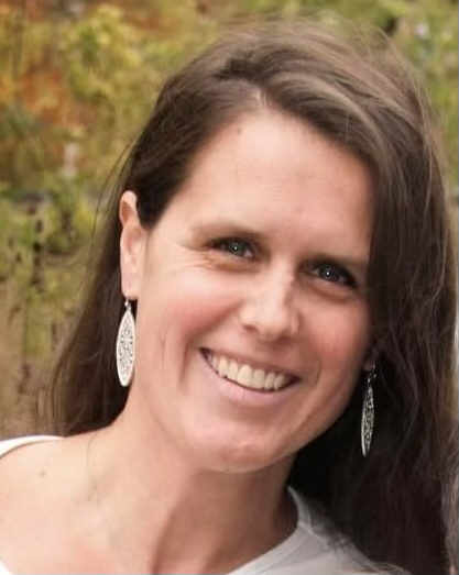 Smiling woman with long dark hair wearing hoop earrings against blurred outdoor background.