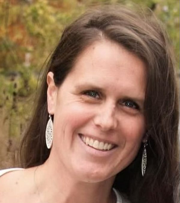 Woman smiling at camera wearing pearl earrings against blurred green foliage background.