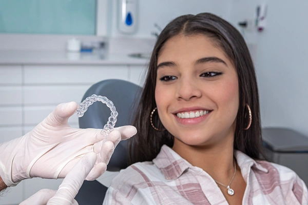 Female orthodontist holding dental aligner in clinical office setting, smiling at camera.