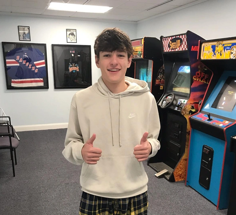 Young man giving thumbs up in arcade game room with posters and arcade machines.